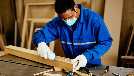 A worker wearing a blue uniform and a mask is applying Fokis wood adhesive onto wood, preparing to bond the wood pieces together.